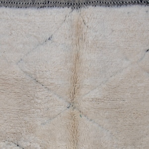 May include: Close-up of a cream-colored, textured rug with a diamond pattern. The rug has a soft, plush appearance with a dark gray border. The design features intersecting lines creating diamond shapes.