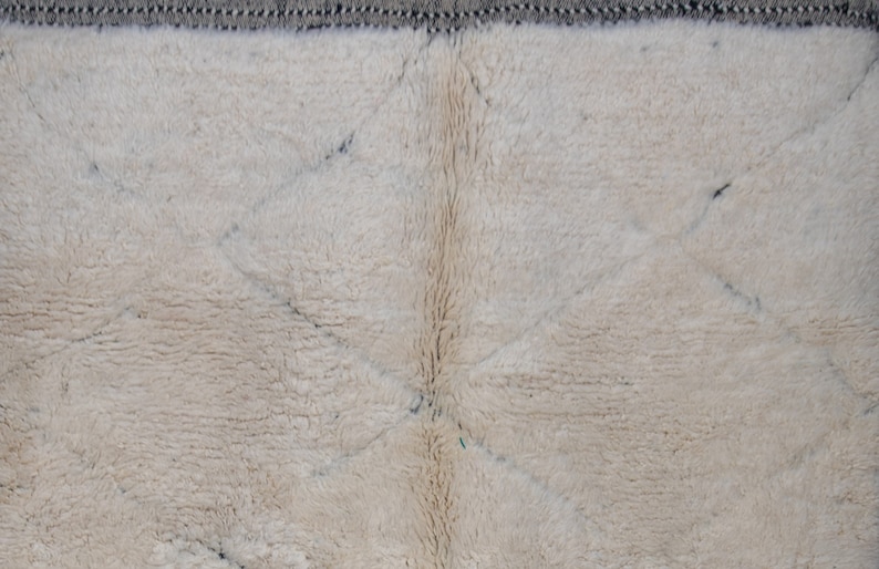May include: Close-up of a cream-colored, textured rug with a diamond pattern. The rug has a soft, plush appearance with a dark gray border. The design features intersecting lines creating diamond shapes.