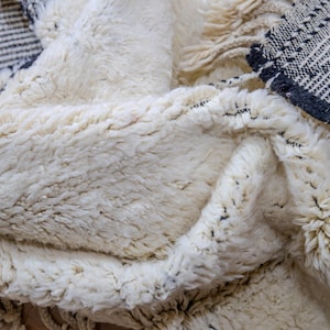 May include: Close-up of a cream-colored, textured rug with a thick pile. The rug has a geometric pattern in black and white along the edges, with fringe detailing. The rug appears soft and inviting.