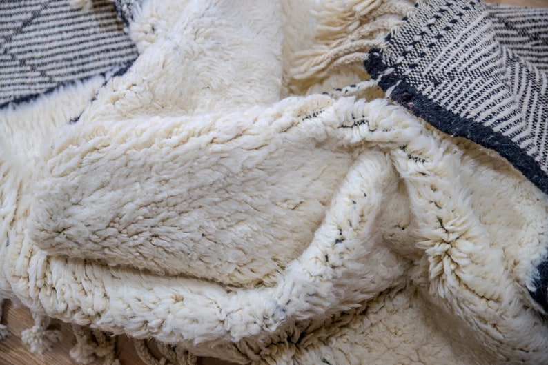 May include: Close-up of a cream-colored, textured rug with a thick pile. The rug has a geometric pattern in black and white along the edges, with fringe detailing. The rug appears soft and inviting.