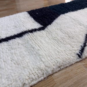 Pu&ograve; includere: Un tappeto geometrico bianco e nero con una texture soffice. Il tappeto ha un design moderno con linee nere su sfondo bianco.