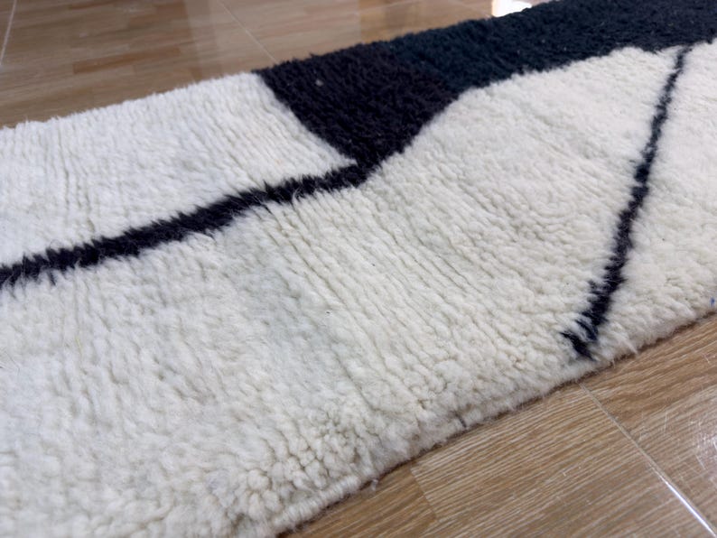 Pu&ograve; includere: Un tappeto geometrico bianco e nero con una texture soffice. Il tappeto ha un design moderno con linee nere su sfondo bianco.