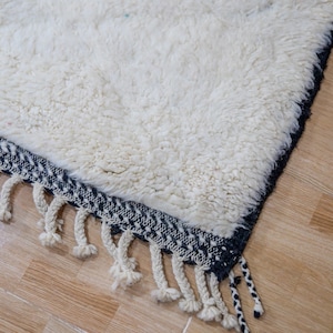 May include: Close-up of a white, textured rug with a dark blue border and cream-colored fringe. The rug has a thick, plush pile and a geometric pattern on the border. The fringe hangs down from the edge of the rug.