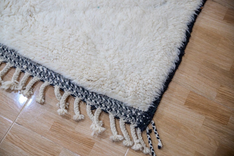 May include: Close-up of a white, textured rug with a dark blue border and cream-colored fringe. The rug has a thick, plush pile and a geometric pattern on the border. The fringe hangs down from the edge of the rug.