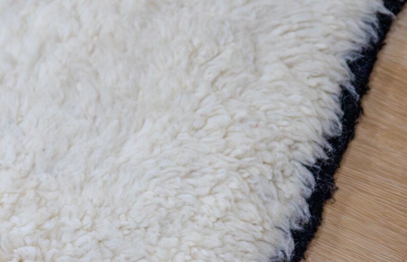 May include: Close-up of a white, fluffy rug with a black border. The rug has a thick, textured pile, creating a soft and inviting appearance. The contrasting black border adds a modern touch to the design.