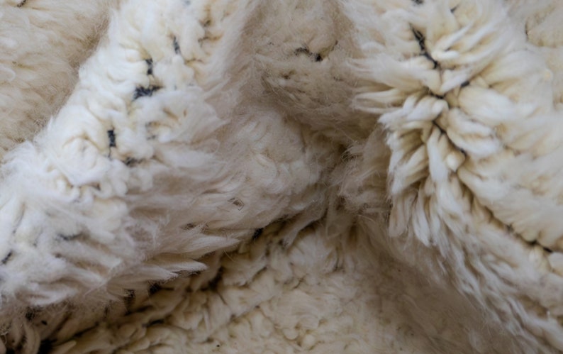 May include: Close-up of a cream-colored, fluffy rug with a thick pile. The rug has a textured surface with small, dark flecks scattered throughout. The fibers are soft and appear to be made of wool.