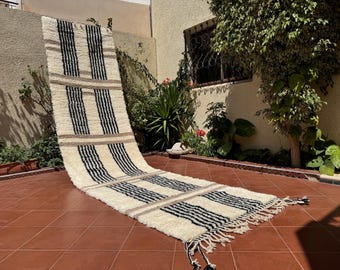 May include: A cream-colored, rectangular rug with vertical black stripes and tan horizontal bands. The rug has a fringed edge and is displayed outdoors on a terracotta tile surface. The rug is made of a thick, textured material.