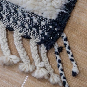 May include: Close-up of a section of a woven rug with a geometric pattern in black and white. The rug has a textured, shaggy appearance with braided fringe in cream and black. The rug is placed on a light brown wooden floor.