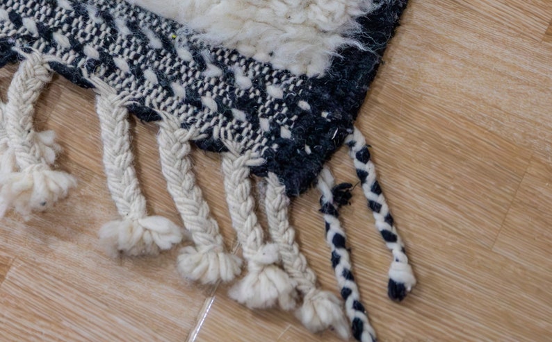 May include: Close-up of a section of a woven rug with a geometric pattern in black and white. The rug has a textured, shaggy appearance with braided fringe in cream and black. The rug is placed on a light brown wooden floor.