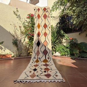 Op de afbeelding: Een crèmekleurig, rechthoekig tapijt met een geometrisch ruitpatroon in tinten oranje, bruin en zwart. Het tapijt wordt buiten tentoongesteld en toont zijn ontwerp en textuur. Het tapijt heeft een franje aan de onderkant.