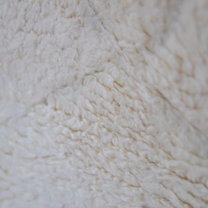May include: Close-up of a cream-colored, textured fabric. The surface has a soft, fluffy appearance with visible fibers and subtle variations in tone. The texture suggests a warm, cozy material.