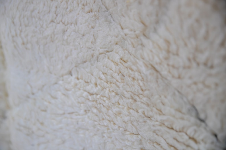May include: Close-up of a cream-colored, textured fabric. The surface has a soft, fluffy appearance with visible fibers and subtle variations in tone. The texture suggests a warm, cozy material.