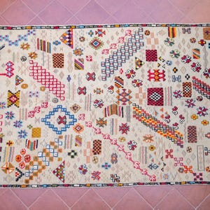 May include: A white rug with a geometric pattern in shades of red, blue, yellow, and orange. The pattern is made up of various shapes, including squares, triangles, and diamonds.