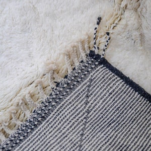 May include: Close-up of a white, textured rug with a shaggy pile. The corner of the rug is folded over, revealing a contrasting black and white striped pattern on the underside. The rug has fringe detailing.