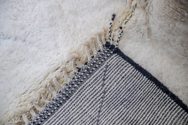 May include: Close-up of a white, textured rug with a shaggy pile. The corner of the rug is folded over, revealing a contrasting black and white striped pattern on the underside. The rug has fringe detailing.