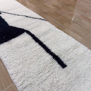 Pu&ograve; includere: Un tappeto di forma allungata e stretta con un motivo geometrico bianco e nero. Il tappeto ha una superficie soffice e testurizzata.