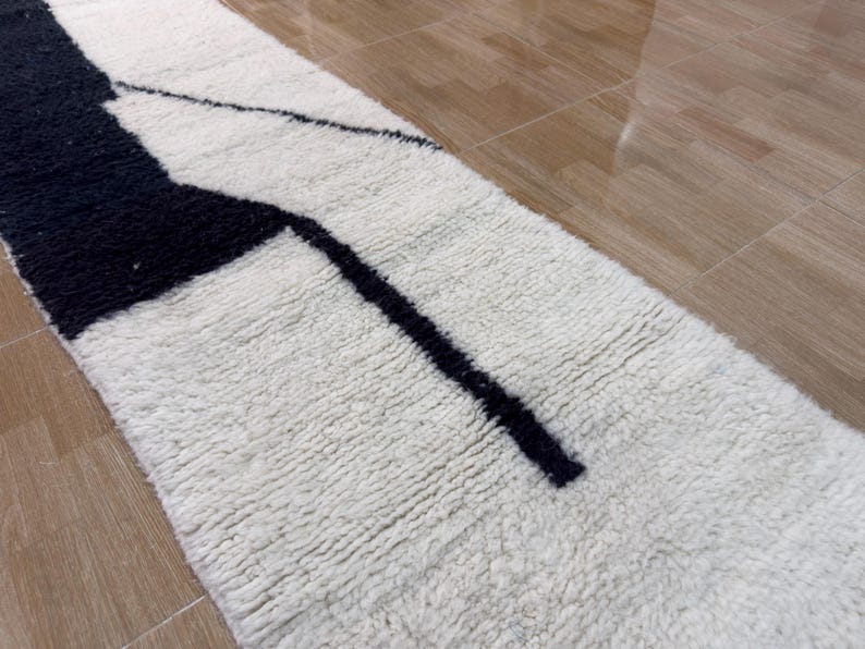Pu&ograve; includere: Un tappeto di forma allungata e stretta con un motivo geometrico bianco e nero. Il tappeto ha una superficie soffice e testurizzata.