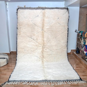 May include: A large, rectangular, off-white rug with a thick, textured pile. The rug has a dark border with fringe along the bottom edge. The rug is displayed in a room with other rugs and cushions.