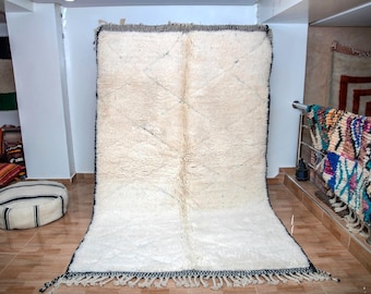 May include: A large, rectangular, off-white rug with a thick, textured pile. The rug has a dark border with fringe along the bottom edge. The rug is displayed in a room with other rugs and cushions.