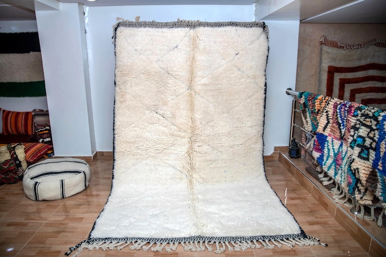 May include: A large, rectangular, off-white rug with a thick, textured pile. The rug has a dark border with fringe along the bottom edge. The rug is displayed in a room with other rugs and cushions.