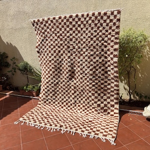 May include: A large, rectangular rug with a checkered pattern in shades of brown and cream. The rug has a thick pile and a fringe along the bottom edge. The rug is displayed outdoors, leaning against a wall.
