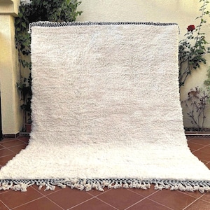 May include: A white shag rug with a black and white fringe border. The rug is laid out on a tiled floor.