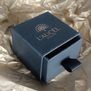 May include: A black box with a gold logo that says "L'AU&Ccedil;EL BIJOUTERIE" and a peacock design. The box has a drawer with a black ribbon pull.