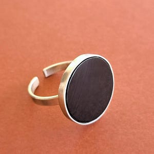 Peut inclure: Bague réglable argentée avec une grosse pierre noire circulaire. La bague a un design minimaliste avec une finition polie. La pierre noire est sertie dans une lunette argentée, créant un contraste saisissant.