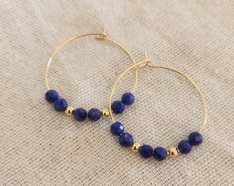 May include: Gold-tone hoop earrings with faceted, deep blue beads and small gold beads. The earrings are displayed on a neutral-colored, textured surface. These earrings are a stylish accessory.