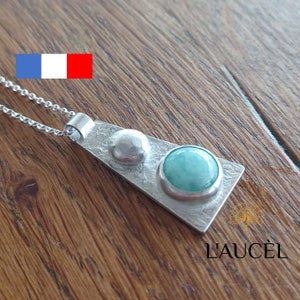 Peut inclure: Un collier avec un pendentif en argent de forme triangulaire et deux pierres rondes. La plus grande pierre est de couleur vert clair et la plus petite est en argent. Le collier est sur une surface en bois.