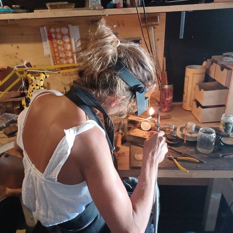 May include: A person wearing a white tank top and a black apron is working on a jewellery project. They are using a torch to heat a piece of metal. The person is wearing safety glasses and a magnifying glass to protect their eyes.