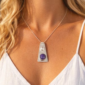 Peut inclure: Un collier pendentif argenté avec un design géométrique. Le pendentif présente une grosse pierre précieuse ronde et violette sertie dans un cadre argenté, avec une petite sphère argentée au-dessus. Le collier est porté sur un haut blanc.