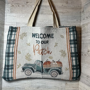 May include: Tote bag with a fall theme. The bag features a design with a vintage truck carrying pumpkins, the text "WELCOME TO OUR PATCH", and plaid accents in green and white. The bag has beige handles.