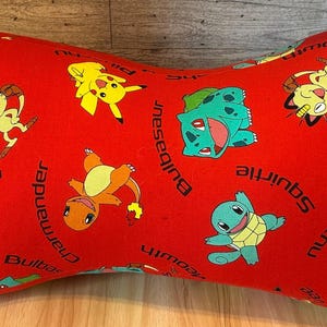 May include: Red, bone-shaped pillow featuring various Pokémon characters like Pikachu, Bulbasaur, and Squirtle. The pillow has a repeating pattern of the characters and their names in black text. A yellow ribbon is attached.