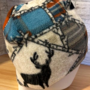 May include: A fleece beanie with a patchwork design in shades of blue, orange, gray, and cream. The beanie features a black silhouette of a deer on a cream-colored section. The hat is designed to keep the head warm.