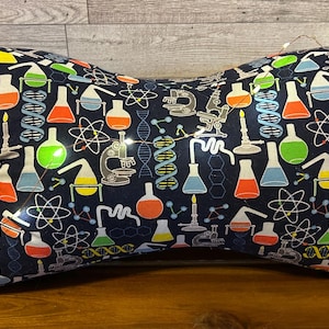 May include: A navy blue, bone-shaped pillow with a science-themed pattern. The pattern includes colorful beakers, microscopes, and atomic symbols. The pillow has a small orange bow and is illuminated with fairy lights.