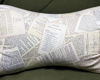 May include: A bone-shaped pillow featuring an off-white fabric cover printed with a collage of vintage book pages, including visible black text, chapter headings, and decorative flourishes arranged in an overlapping, layered pattern.