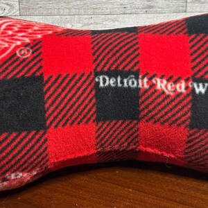 May include: Bone-shaped pillow with a red and black buffalo plaid pattern. The pillow features the Detroit Red Wings logo and text in white. The pillow is made of a soft, plush material.
