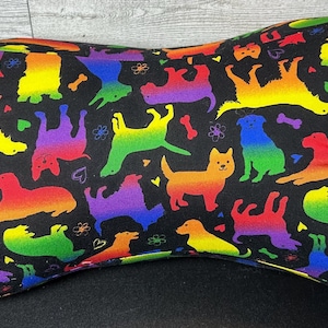 May include: A bone-shaped pillow featuring a black fabric background with a repeating pattern of rainbow-colored dog silhouettes, small hearts, flowers, and bone shapes. The pillow has small red fabric loops on each end for hanging or carrying.