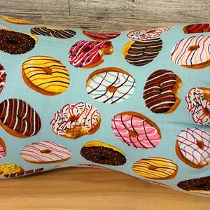 May include: Bone-shaped pillow with a light blue background covered in colorful donut illustrations. The donuts feature various toppings and colors, including pink, yellow, and chocolate, creating a playful and appetizing design.