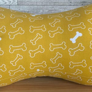 May include: Mustard yellow dog bone-shaped pillow with a repeating pattern of white dog bones. The pillow is made of soft fabric and is designed for comfort and support. The pillow is approximately 20 inches long.