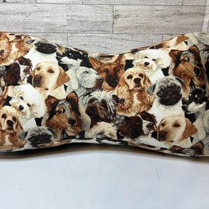 May include: Bone-shaped pillow featuring a collage of various dog breeds in shades of brown, tan, black, and white. The pillow has a cream-colored fabric loop on one side. The background is a light gray and white wood-paneled wall.
