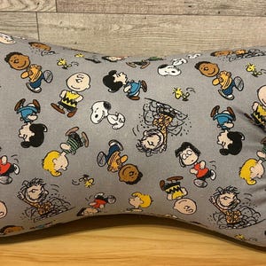 May include: Gray, bone-shaped pillow featuring a repeating pattern of cartoon characters, including Charlie Brown and Snoopy. The pillow has yellow fabric loops on each end. The pillow is designed for neck support.