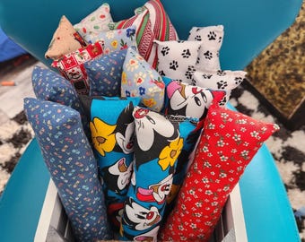 May include: A white wooden crate filled with various cat toys. The toys are in different shapes and patterns, including floral, paw prints, and cartoon characters. The crate has a rope handle and sits on a blue surface.