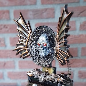 Może przedstawiać: Zegar w stylu steampunk z czaszką i skrzydłami. Zegar ma czarną podstawę ze złotymi akcentami i tarczę zegarową z cyframi rzymskimi. Zegar stoi na drewnianej powierzchni.