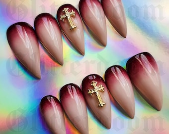 May include: A set of ten stiletto-shaped press-on nails featuring a dark burgundy to nude gradient ombre finish. Two of the nails are adorned with a three-dimensional gold-toned cross charm centered on the nail bed, adding texture to the design.