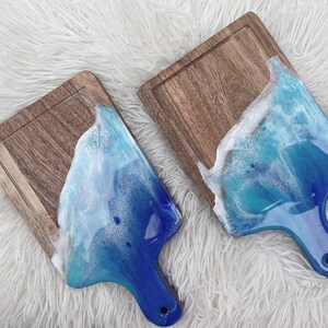 Puede incluir: Dos tablas de cortar de madera con diseños de olas de océano de resina azul y blanca. Las tablas tienen un mango en la parte inferior.
