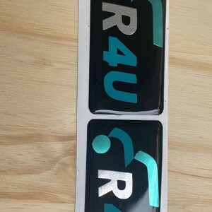 Puede incluir: Dos pegatinas negras y blancas con un logotipo turquesa y plateado que dice "R4U" con un diseño estilizado.