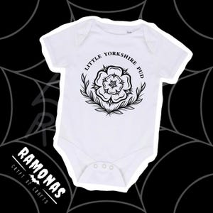 May include: White baby onesie with a black floral design and the text "LITTLE YORKSHIRE PUD" in a circular pattern. The onesie has short sleeves and three snaps at the bottom. The logo "RAMONAS CRYPT OF CRAFTS" is at the bottom left.