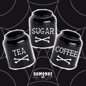 May include: Three black metal canisters with white lettering and bone cross designs. The canisters are labeled "Sugar", "Tea", and "Coffee".